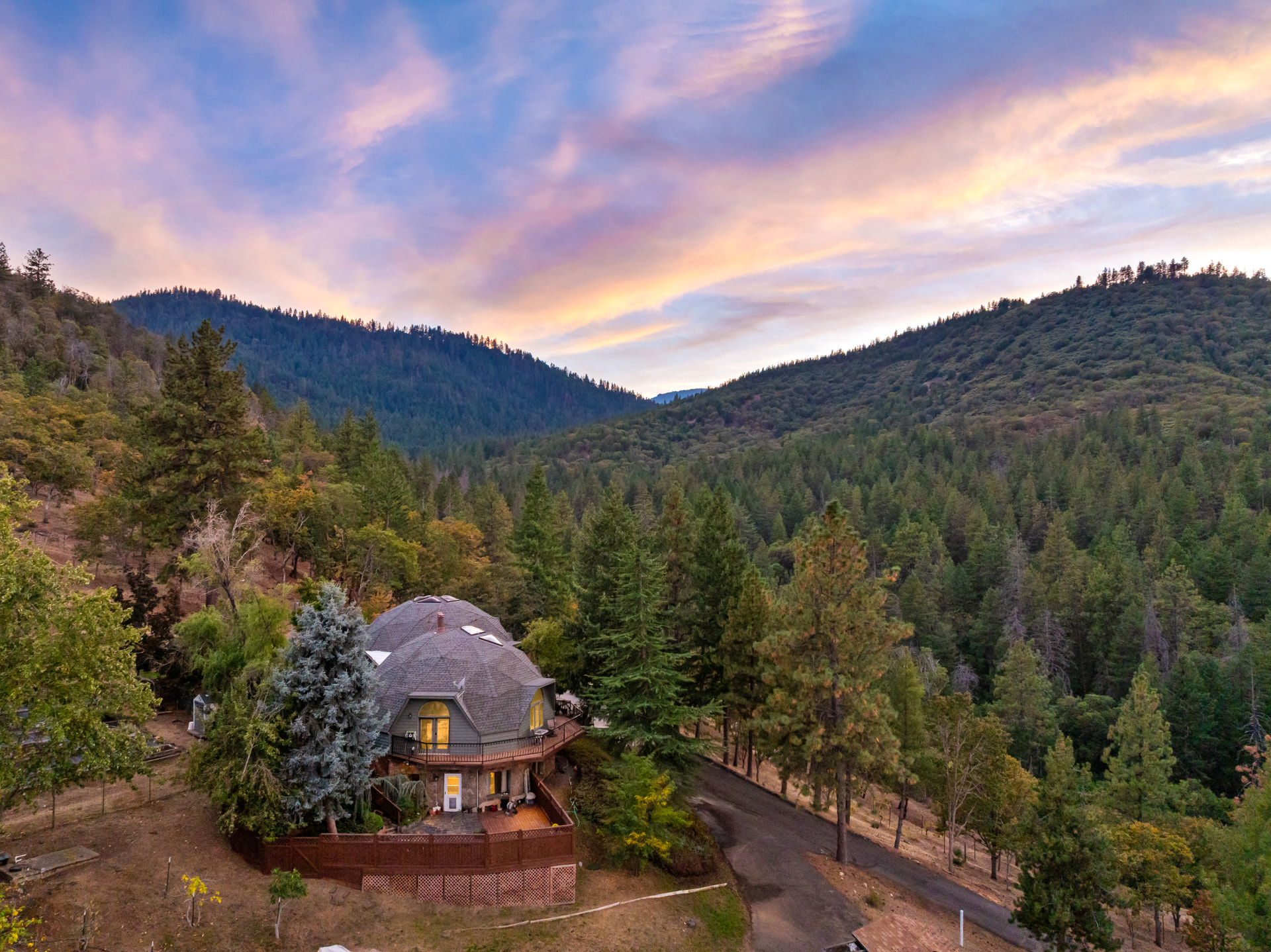 Twilight aerial view of the connected double geodesic dome residence on its traditional foundation, warm light glowing from the wrap-around deck windows, surrounded by conifers on the wooded hillside above the Ashland valley